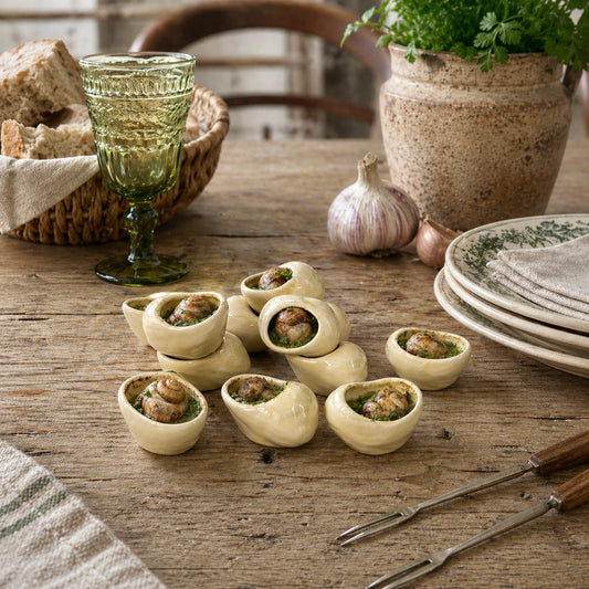 12 small French ceramic escargot snail pots filled, on a wooden table with a green glass, garlic, and a plant.