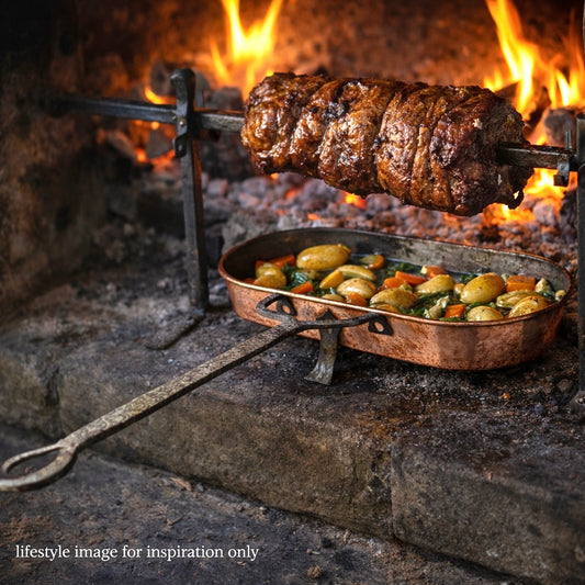 Meat roasting on a spit over an open fire with vegetables in a pan below.