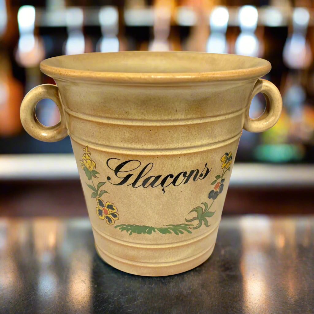 Beige ceramic pot with floral designs and 'Glaçons' text on a bar top with a blurred bar in the background