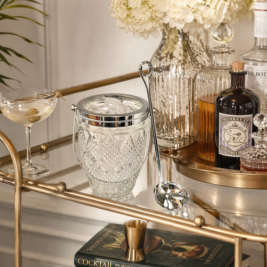 Bar cart with cocktail-making items including a glass ice bucket, bottles, and a cocktail book.