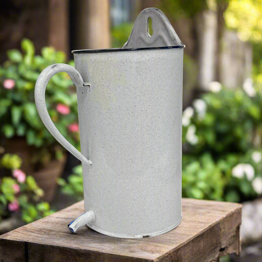 White speckled enamel jug or planter with a spout on a table in a garden with a blurred background of flowers for sale by All Things French Store