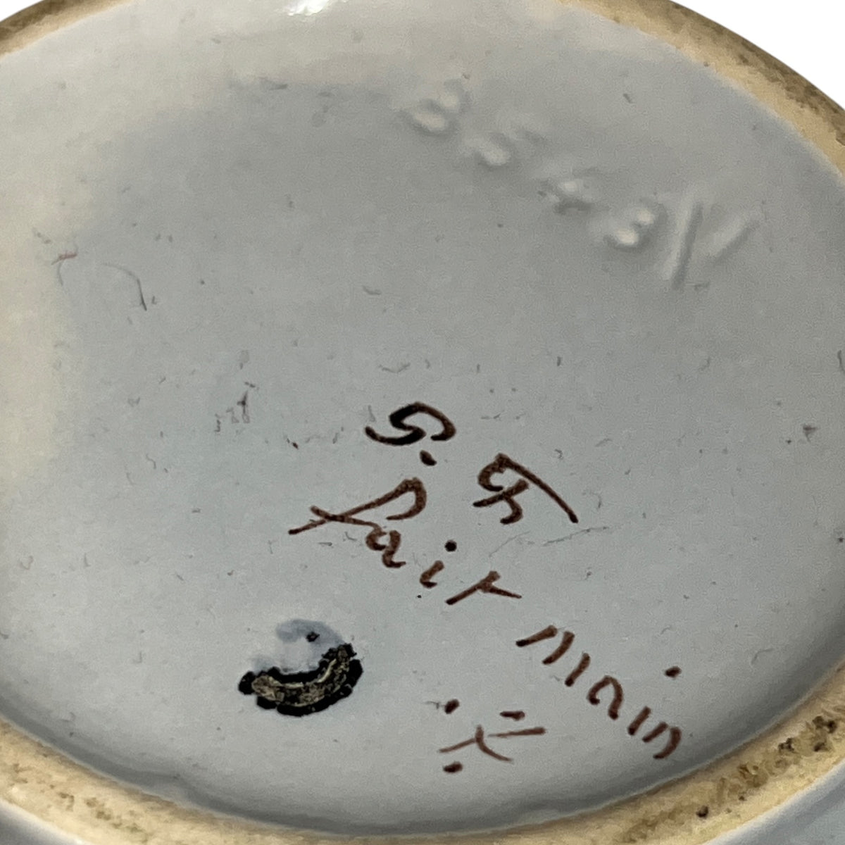 Close-up of the interior bottom of a ceramic dish or bowl, showing a light gray glazed surface with embossed numbering, a small dark stain, and handwritten markings reading 'R.R.
