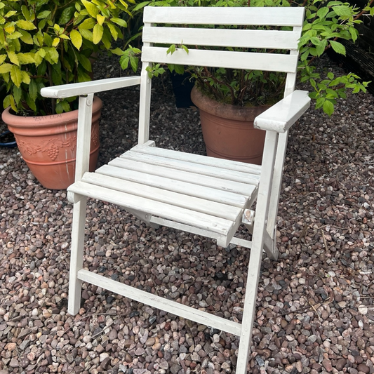 A charming French vintage wooden slatted folding chair set in a picturesque garden with a greenhouse, potted plants, and a gravel path, perfect for adding rustic country cottage charm.