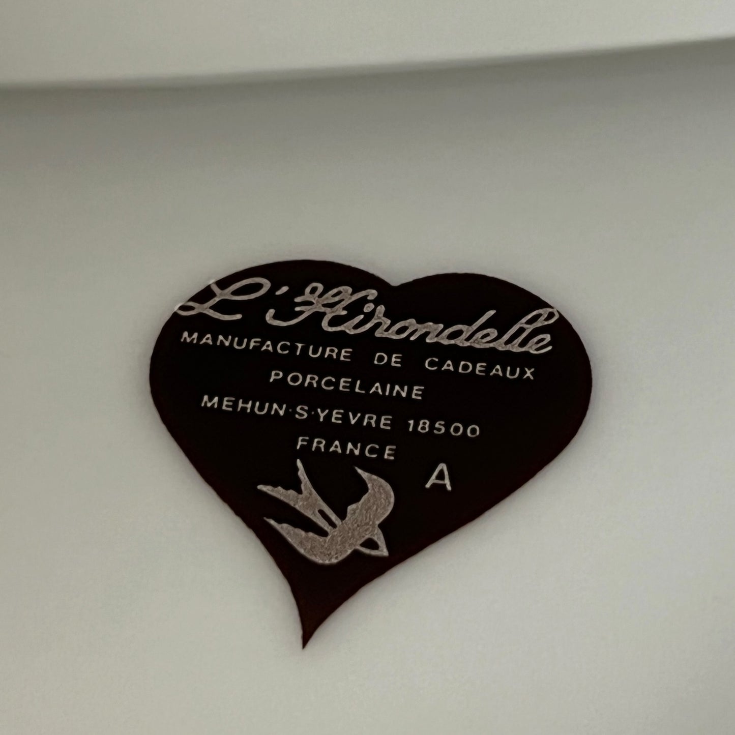A heart-shaped dark brown plaque with white French text reading 'L'Hirondelle Manufacture de Cadeaux Porcelaine' and a silver bird emblem, photographed against a light neutral background.