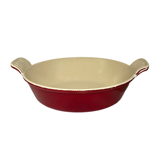 A round red enameled baking dish with a cream-colored interior and dual ribbed side handles, shown against a plain white background.