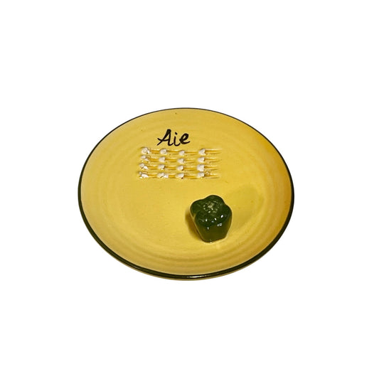 A yellow plate featuring a green pepper and the French word for garlic, “Ail,” designed for grating garlic and perfect for a French farmhouse-style kitchen, set against a white background.