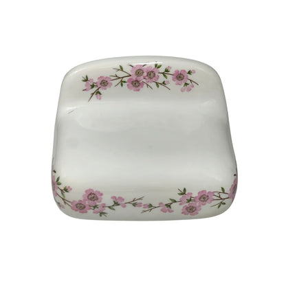 A square white ceramic dish with rounded corners, decorated with delicate pink floral branches along the top and bottom edges, shown against a plain white background.