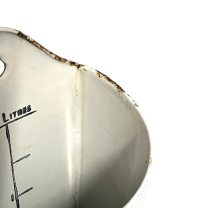 Close-up of the interior of a white metal measuring container with printed litre markings and a worn, rusted rim against a white background.
