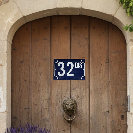 French rectangular vintage enamel door number sign 32 bis with white lettering on a blue background, for an authentic rustic French style front door, set on wooden arched door with a lion shaped knocker , a stone surround and heather and greenery in the corner