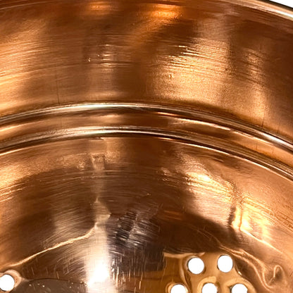 Close-up of the interior of a copper pot or pan, showing a brushed metallic copper surface with a perforated drain hole at the bottom.
