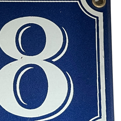 Close-up of a blue house number sign showing the number 8 in white with a metal mounting hole in the top right corner.