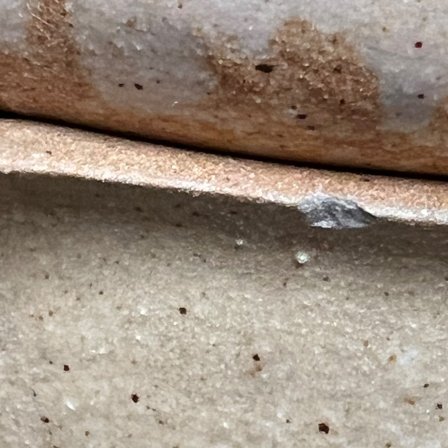 Close-up of a ceramic tile or tile edge showing a textured matte finish with brown speckles and a metallic silver solder joint.