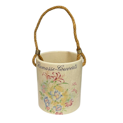A cylindrical ceramic utensil pot in a warm cream colour, decorated with a colourful floral pattern and the French text 'Ramasse-Couverts' in black script. It features a natural jute rope handle wrapped with small metal connectors, set against a plain white background.