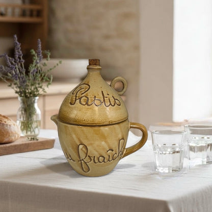 Decorative ceramic jar with 'Pastis' text on a table with glasses and bread.