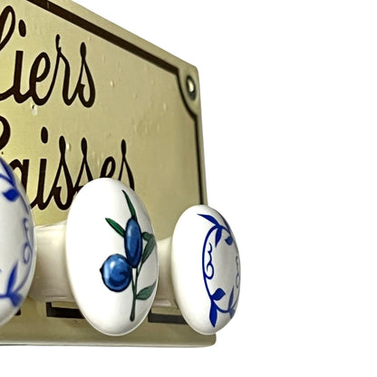 Close-up of a decorative metal sign with three white oval knobs featuring blue and green painted motifs, including blueberries and a fish, set against a white background.
