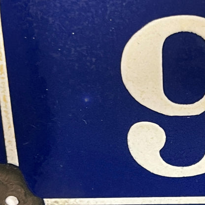 Close-up of a blue enamel-style sign with a large white serif letter or number and a thin white border.