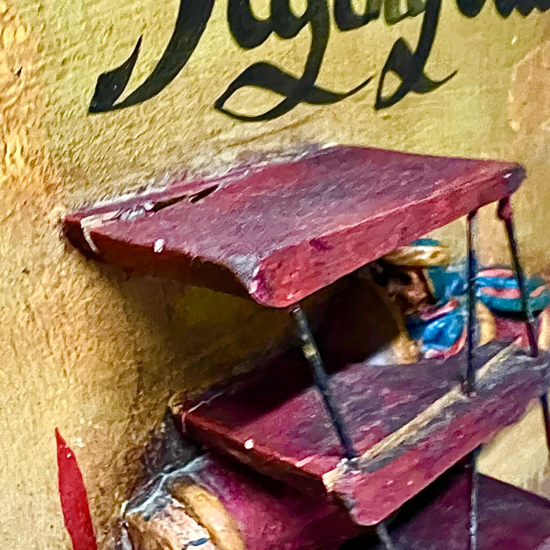 A close-up of a wooden image painted in deep red and burgundy tones, mounted against a textured beige wall with partial black script lettering visible above.