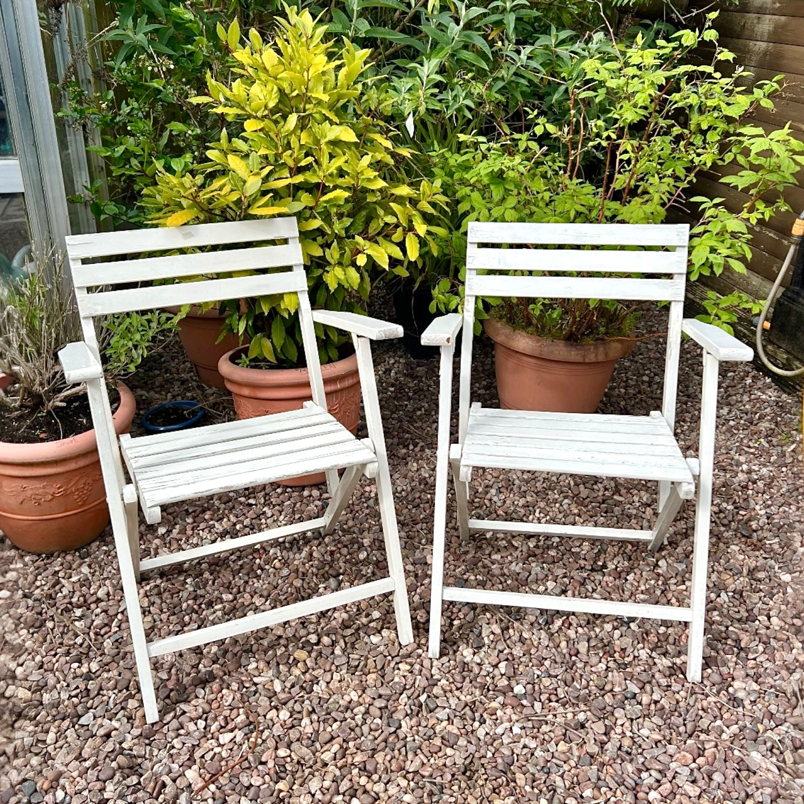 A pair of French vintage wooden slatted garden folding chairs sit in a charming garden setting on a gravel path, perfect for a cosy country cottage garden.