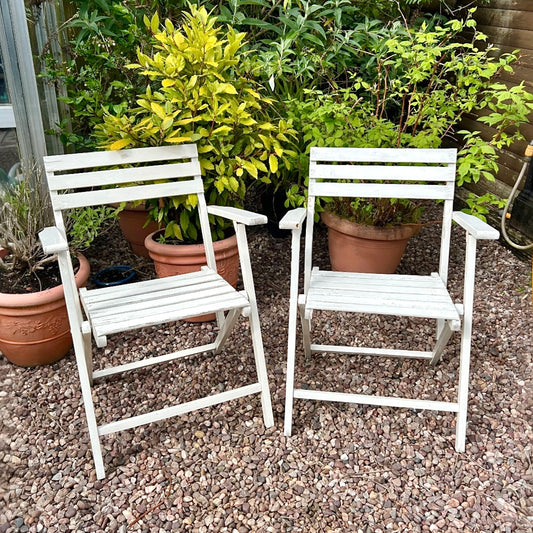 A pair of French vintage wooden slatted garden folding chairs sit in a charming garden setting on a gravel path, perfect for a cosy country cottage garden.