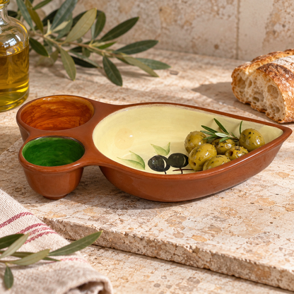 A terracotta-coloured ceramic serving platter with two small dipping bowls, shown on a stone surface with olives, bread, olive branches, and a bottle of olive oil.