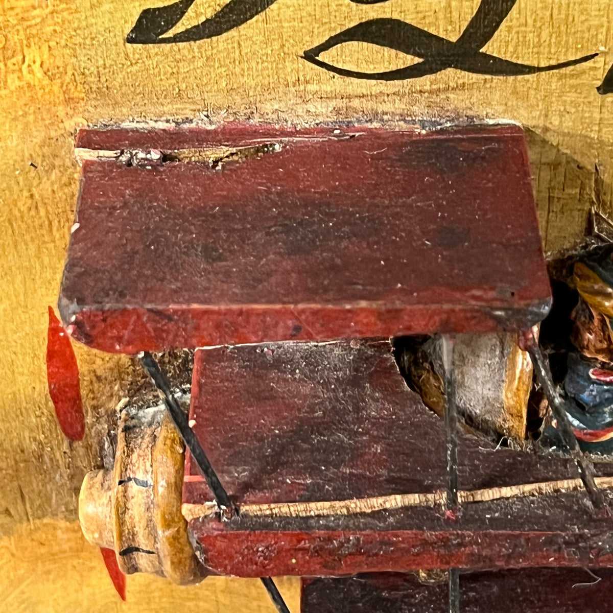Close-up detail of a vintage wooden model airplane with a dark red painted body, showing the wing, fuselage, and part of the propeller against a yellow background with black script lettering.