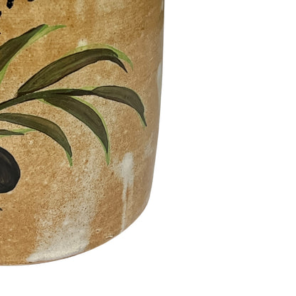 Close-up of a ceramic jar with a textured beige-brown glaze, painted green leaves, and a dark fruit, shown against a plain white background.