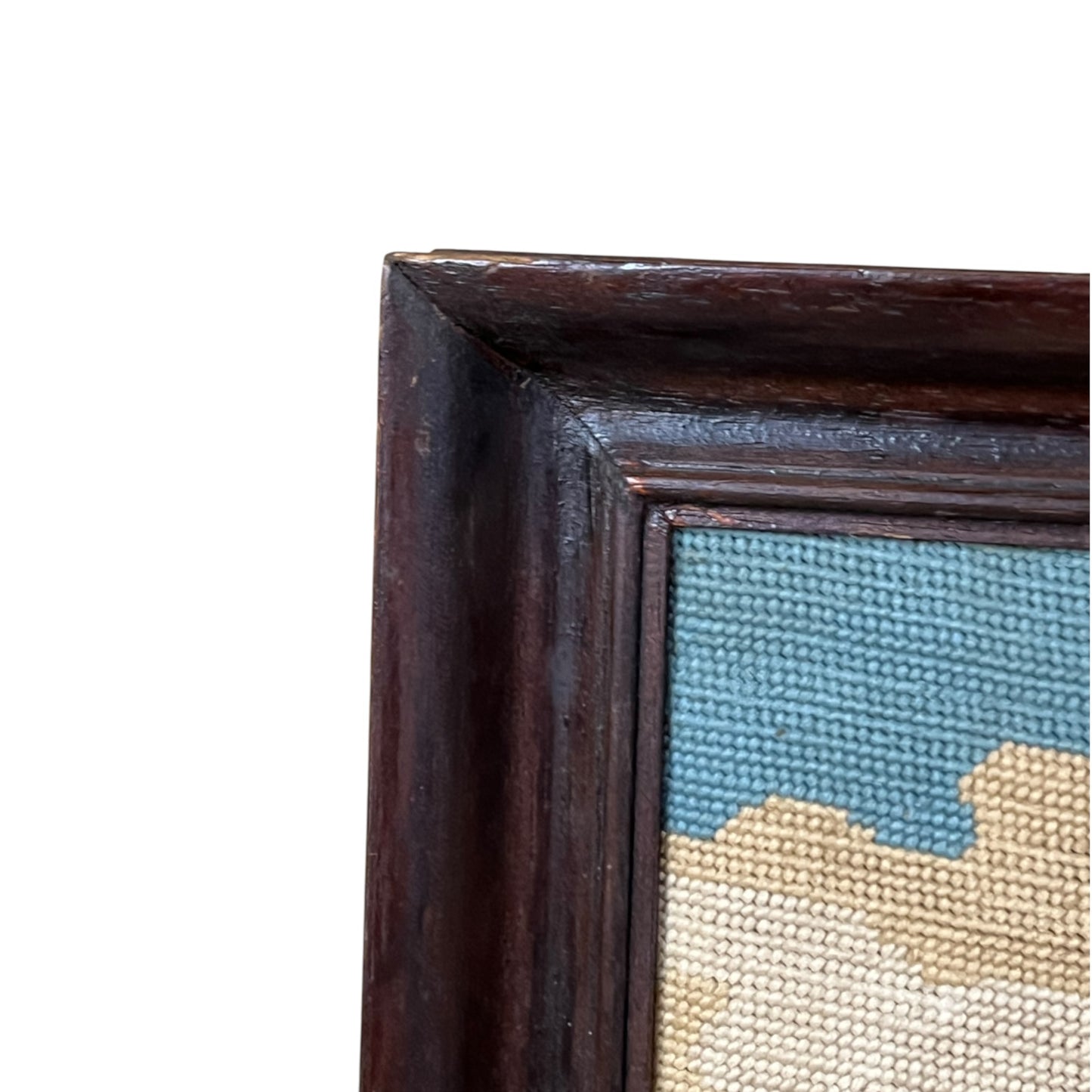 Close-up of the corner of a framed needlepoint canvas, showing a dark brown wooden frame and a blue and beige woven design on a white background.