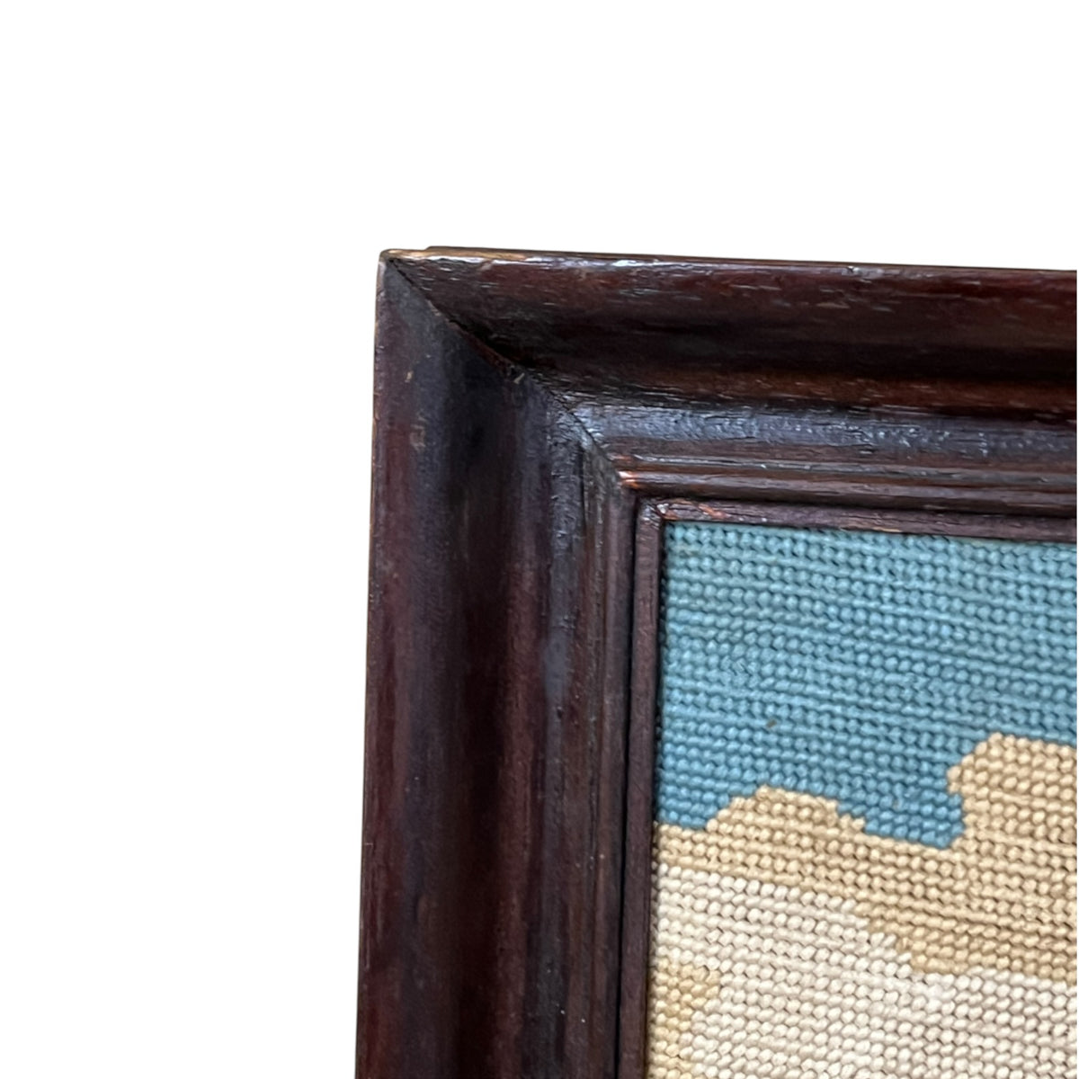 Close-up of the corner of a framed needlepoint canvas, showing a dark brown wooden frame and a blue and beige woven design on a white background.
