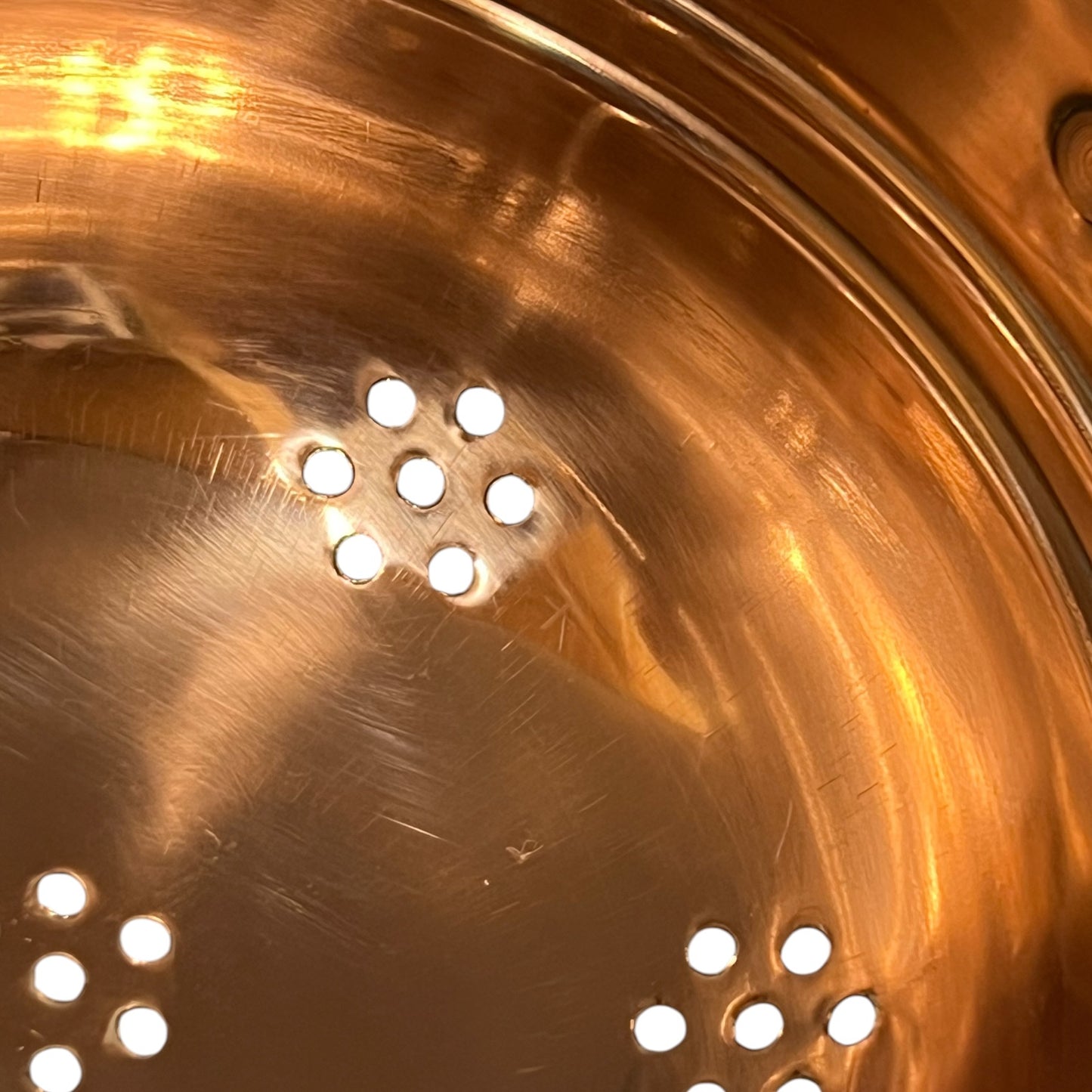 Close-up of the inside of a copper or copper metal pot or pan, showing a brushed metallic surface with circular drainage holes near the bottom.