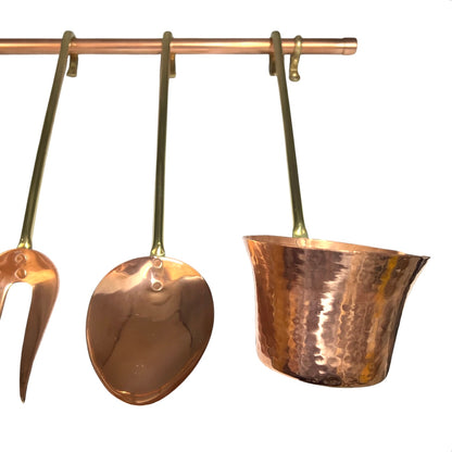 A set of hammered copper kitchen utensils hanging from a copper rod, including a ladle, a fork, and a small pot or cup, shown against a plain white background.