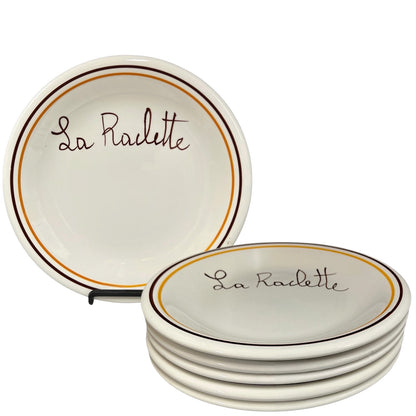 A set of six white ceramic plates with 'La Raclette' written in brown cursive, bordered by a thin gold line and a thicker dark brown line, shown on a white background with one plate standing upright and five stacked.