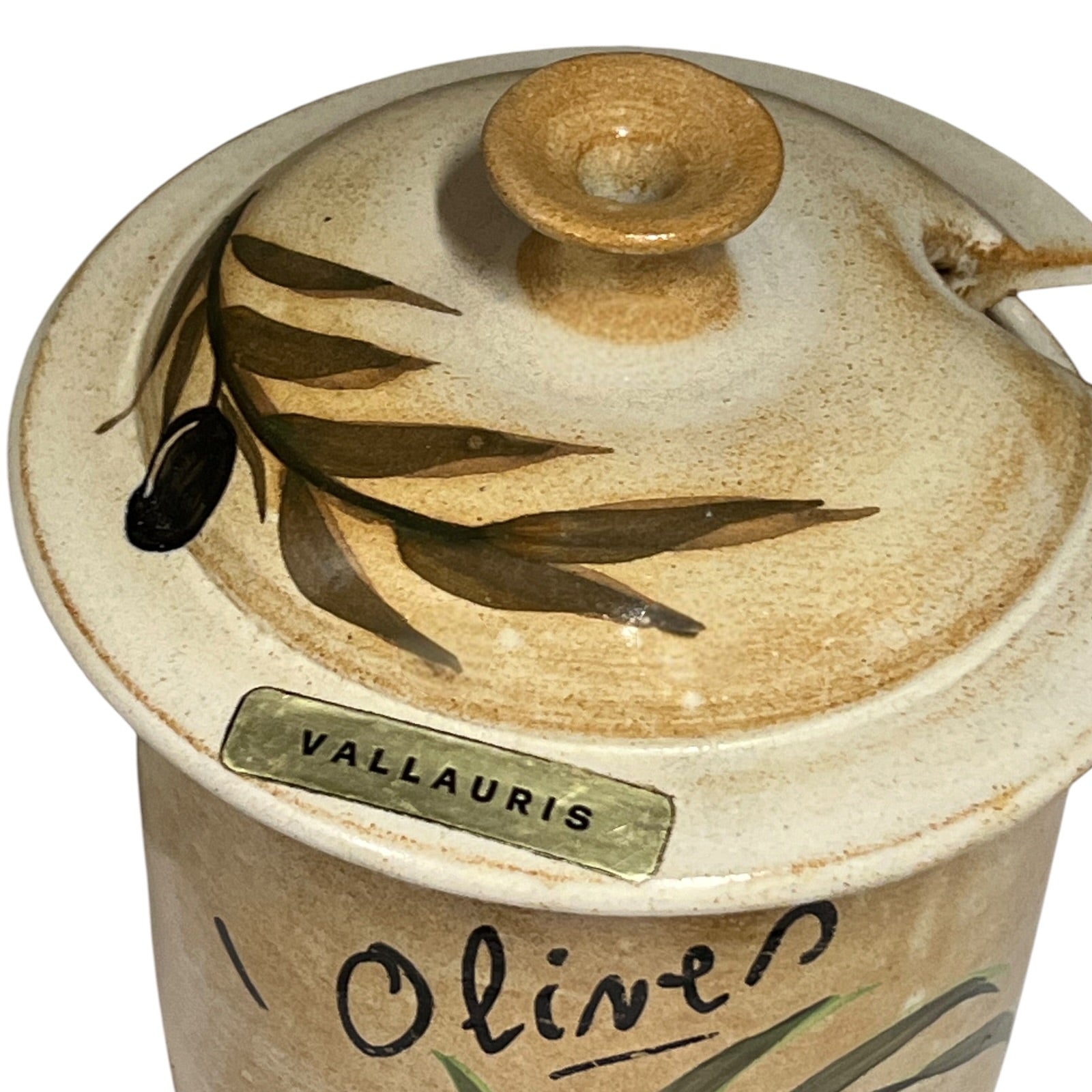A close-up of a handmade ceramic lidded container or jar with a rustic beige glaze, decorated with olive branch motifs and the word 'Olive' written on the front. A small label reads 'VALLAURIS'.