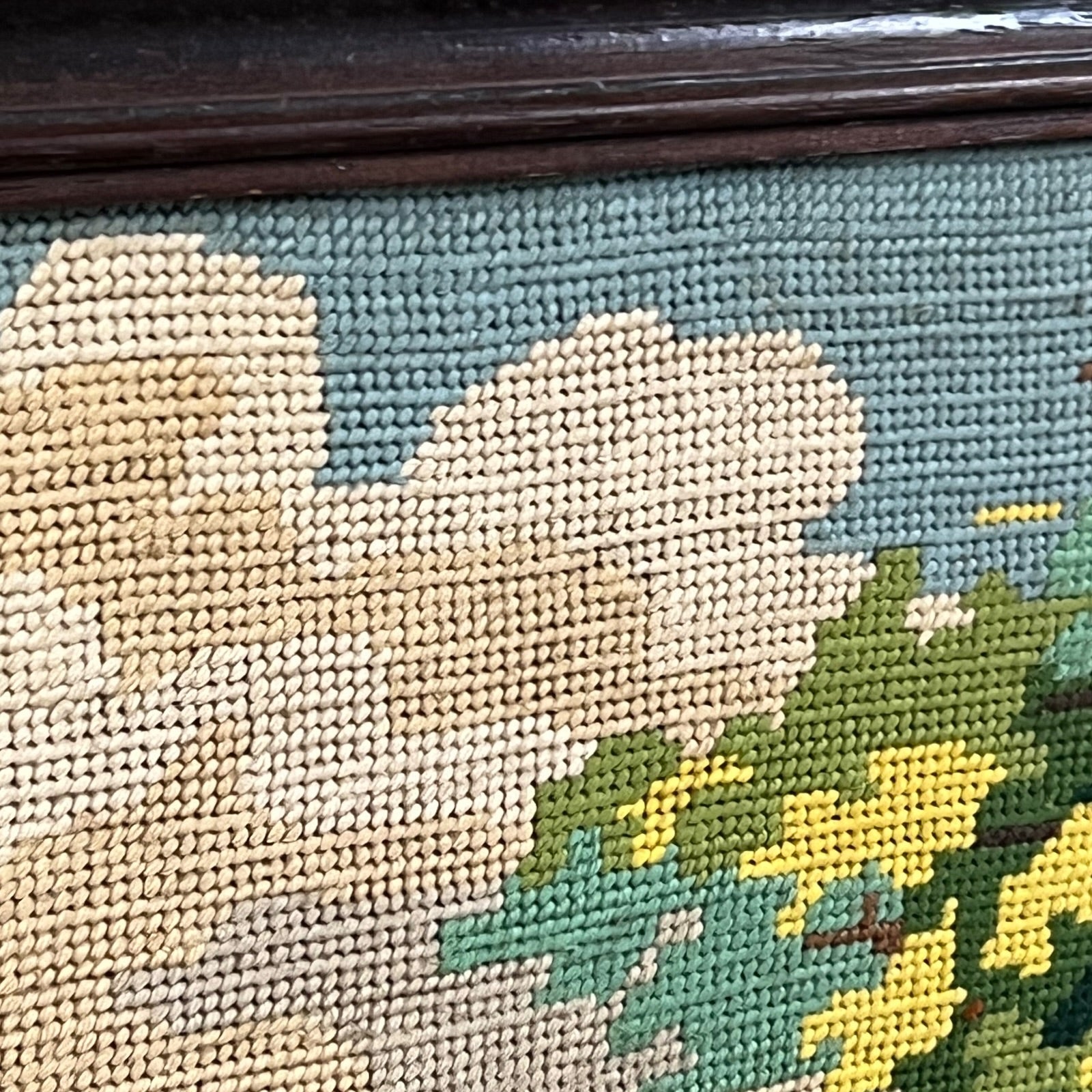Close-up of a needlepoint canvas featuring a landscape design with a beige cloud, green trees, and yellow flowers on a muted blue-green background, framed by a dark wooden border.