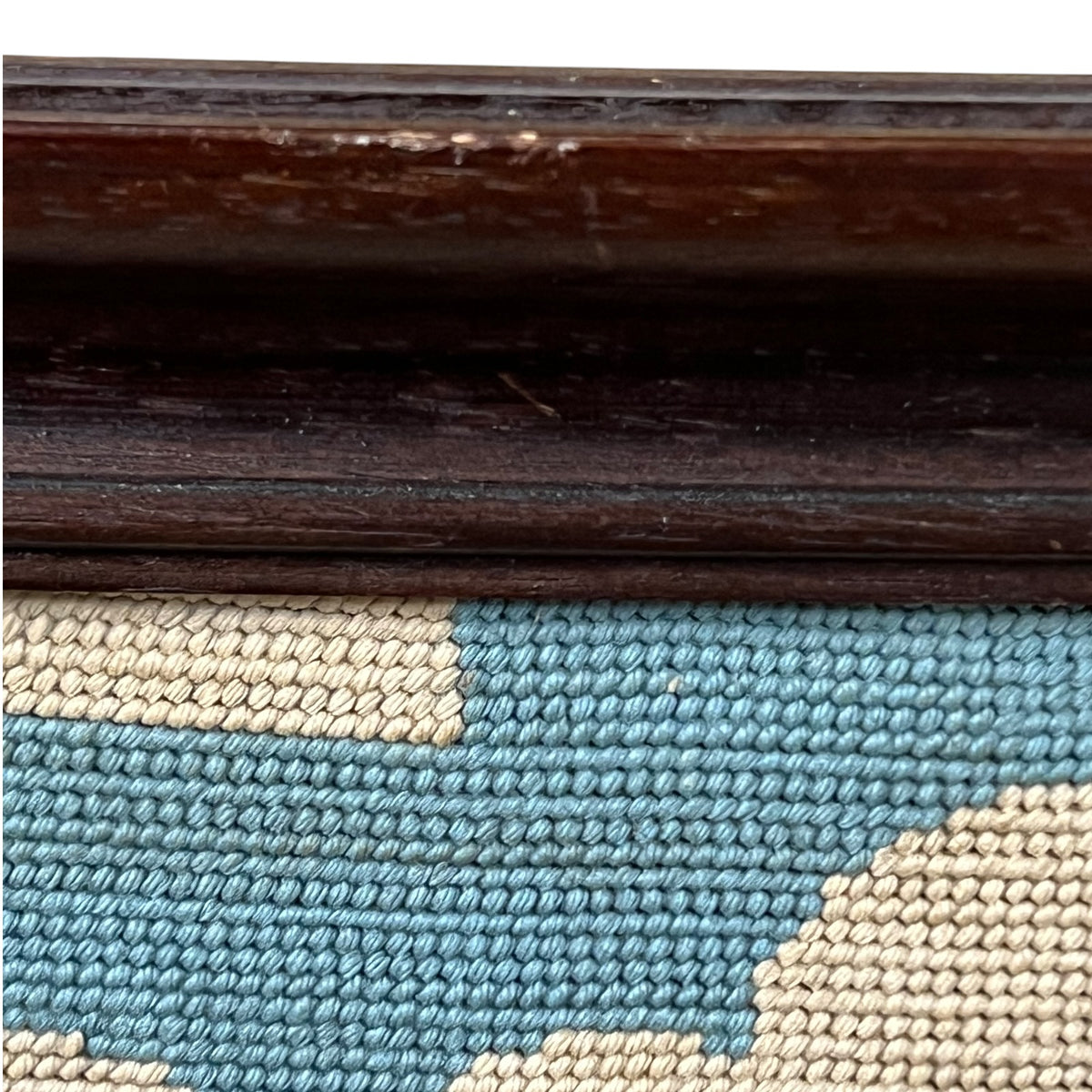 Close-up detail of a framed needlepoint canvas showing a dark brown wooden frame and a blue and beige patterned needlepoint surface.