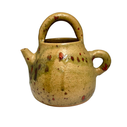 A small handmade ceramic teapot with a rounded body, loop handle, and small side spout, covered in a speckled beige and olive glaze with scattered reddish-brown spots, isolated on a white background.