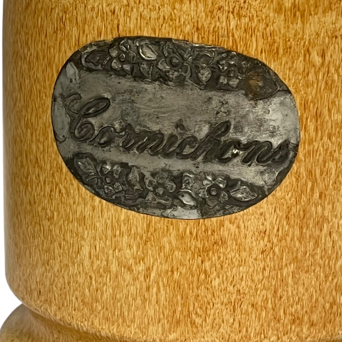 Close-up of a cylindrical container or mug with a round metal label reading 'Cornichons' in cursive, surrounded by floral decoration, on a warm tan wood-grain surface.