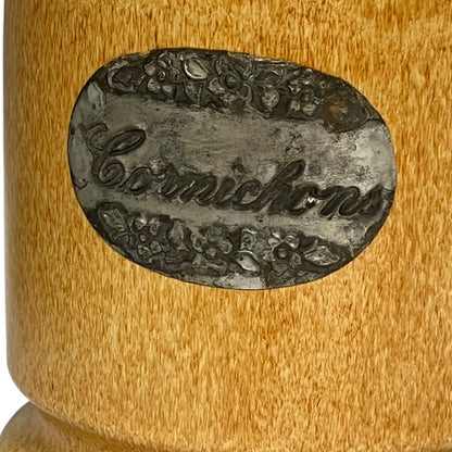 Close-up of a cylindrical container or mug with a round metal label reading 'Cornichons' in cursive, surrounded by floral decoration, on a warm tan wood-grain surface.