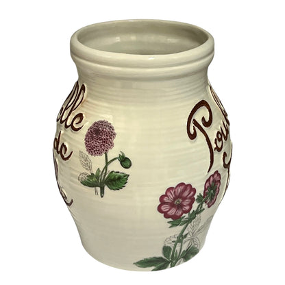 A cream-coloured ceramic jar with a ribbed texture, featuring hand-painted floral motifs in purple, green, and white, along with brown cursive text, set against a plain white background.