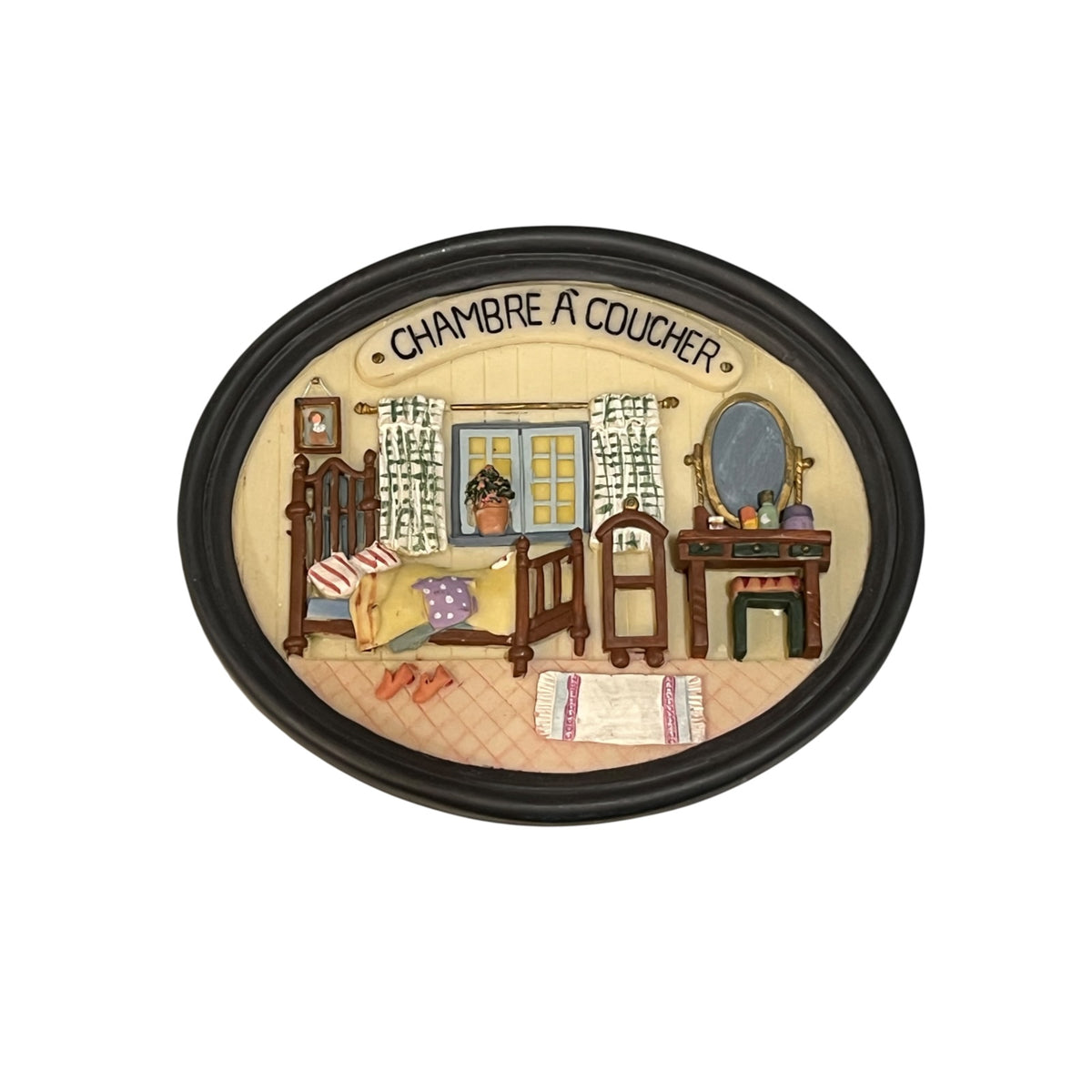 An oval decorative plaque or sign with a black frame and the French text 'CHAMBRE A COUCHER' at the top. The center shows a painted bedroom scene with a bed, dresser, mirror, curtains, window, and small furnishings on a cream background.