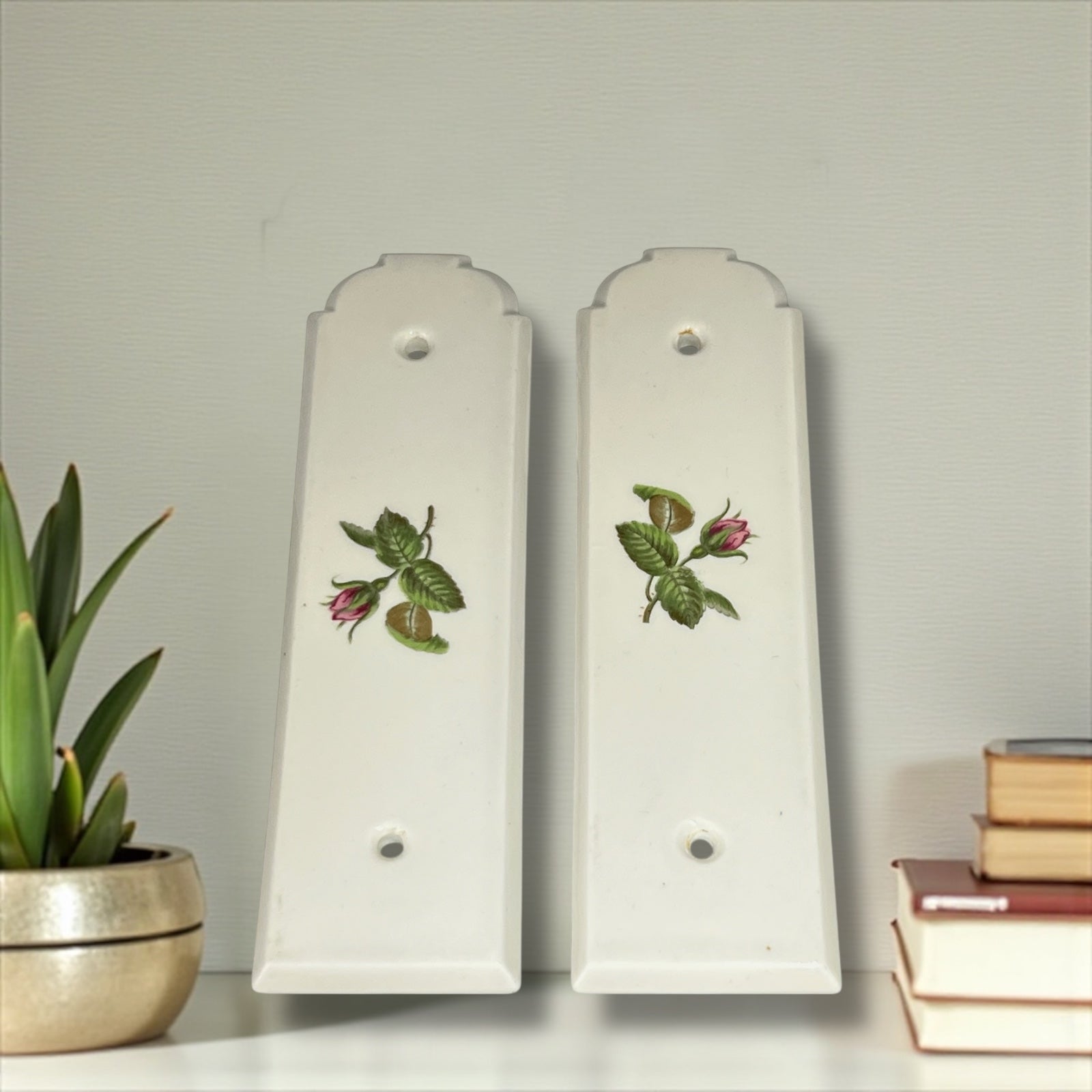 Two white finger door push plates with floral designs on a shelf with a plant, and books on a light grey background.