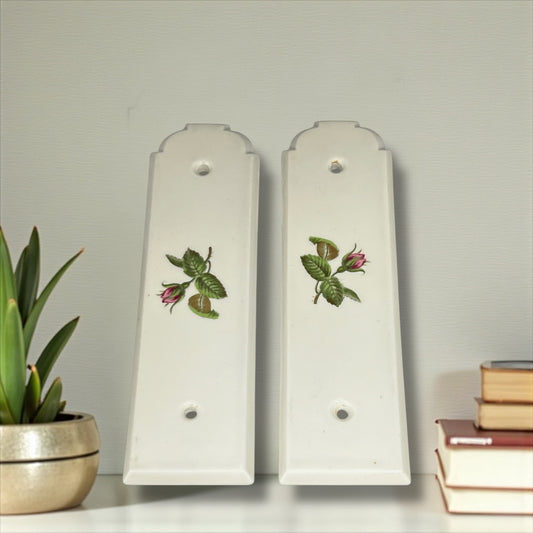 Two white finger door push plates with floral designs on a shelf with a plant, and books on a light grey background.