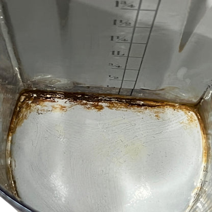 Close-up of the inside of an enamel measuring pot or saucepan, showing a heavily stained white surface with brown rust and grime along the bottom edge and upper side walls.