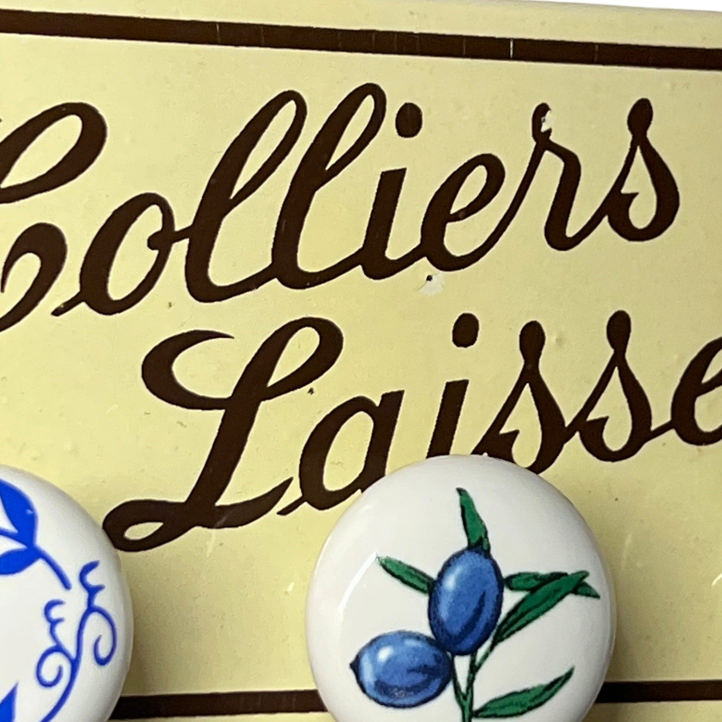 Close-up of a cream-colored sign with dark brown script reading 'Colliers Laisse' and two round white decorative buttons or medallions, one with blue floral detailing and the other with blue olives on green stems.