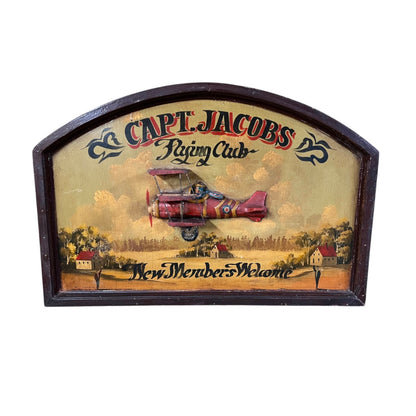 A vintage wooden sign for 'Capt. Jacobs Flying Club' featuring an illustration of a red biplane flying over a small town scene.