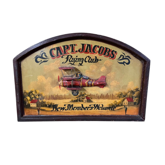 A vintage wooden sign for 'Capt. Jacobs Flying Club' featuring an illustration of a red biplane flying over a small town scene.