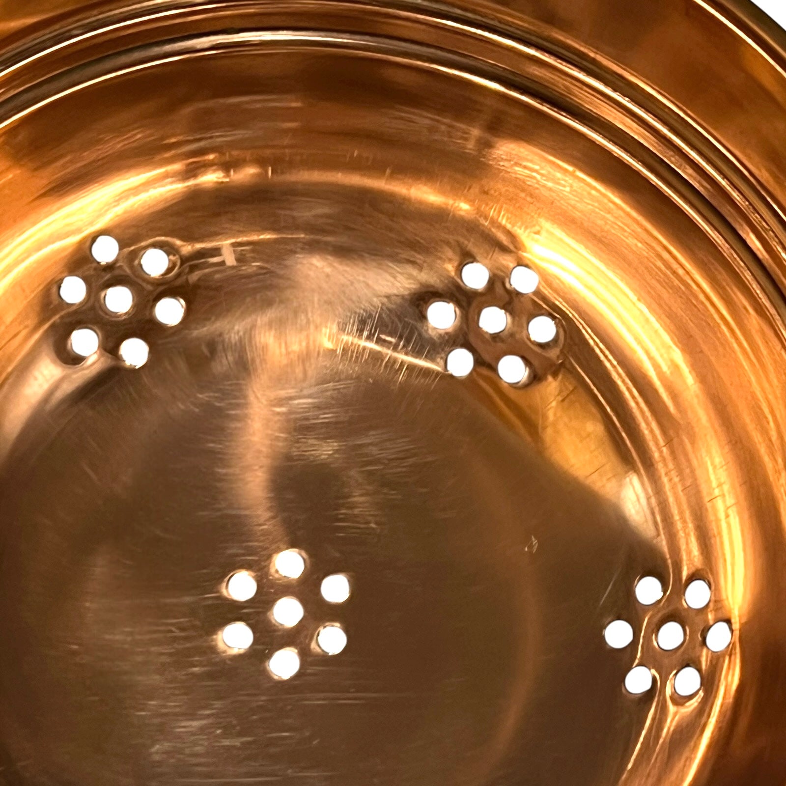 Close-up of the inside of a metallic copper or bronze pot, showing a brushed metal surface with several small circular holes arranged near the rim and one near the bottom.