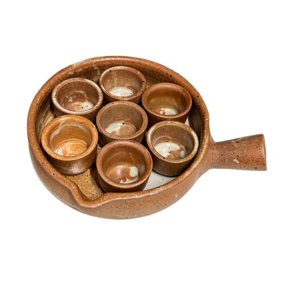 A handmade brown ceramic set of six small cups arranged inside a larger shallow bowl with a handle, photographed on a plain white background.