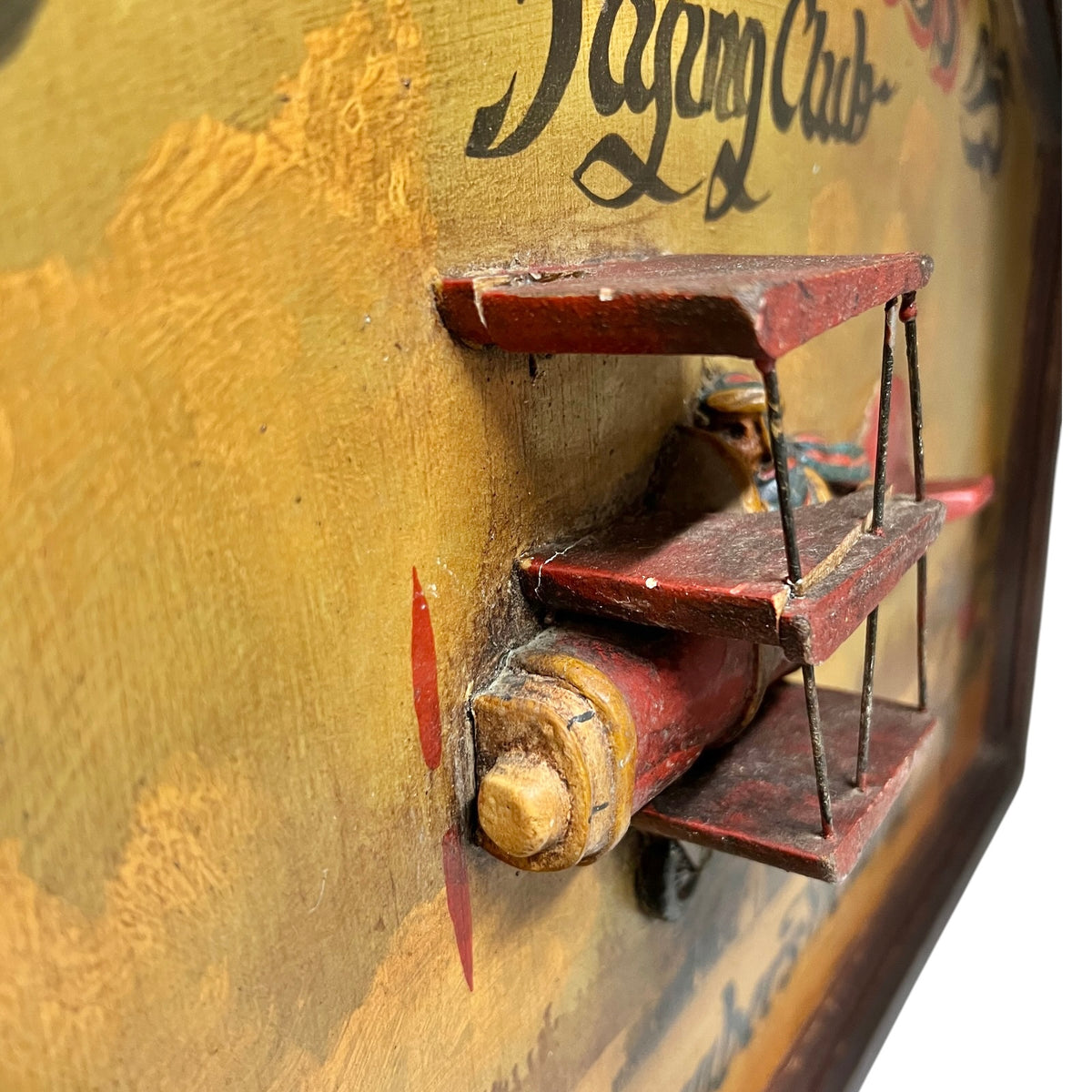 Close-up of a vintage wooden picture or wall decor piece with a distressed yellow painted surface, black script lettering, and a small red-brown wooden shelf or bracket mounted on the front.