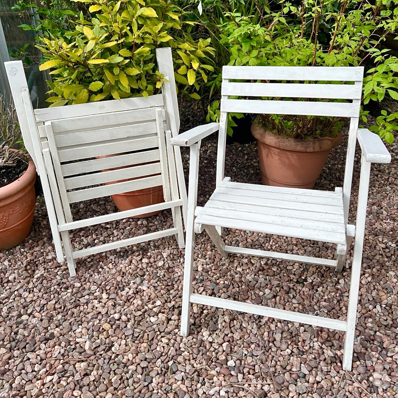 A pair of French vintage wooden slatted garden folding chairs sit in a charming garden setting on a gravel path, perfect for a cosy country cottage garden.