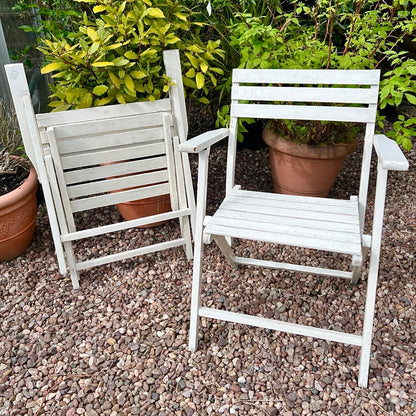 A pair of French vintage wooden slatted garden folding chairs sit in a charming garden setting on a gravel path, perfect for a cosy country cottage garden.